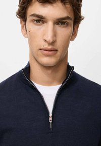Granatowy sweter na zamek błyskawiczny z kołnierzem stójką i srebrnym zamkiem, noszony na białej koszulce. Dzianinowa faktura, dopasowany krój, minimalne zdobienia.
