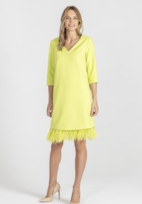 Kobieta w jasnożółtej sukience do kolan z rękawami o długości trzech czwartek i piórkowym dołem, w zestawieniu z nagimi szpilkami, stojąca na białym tle.