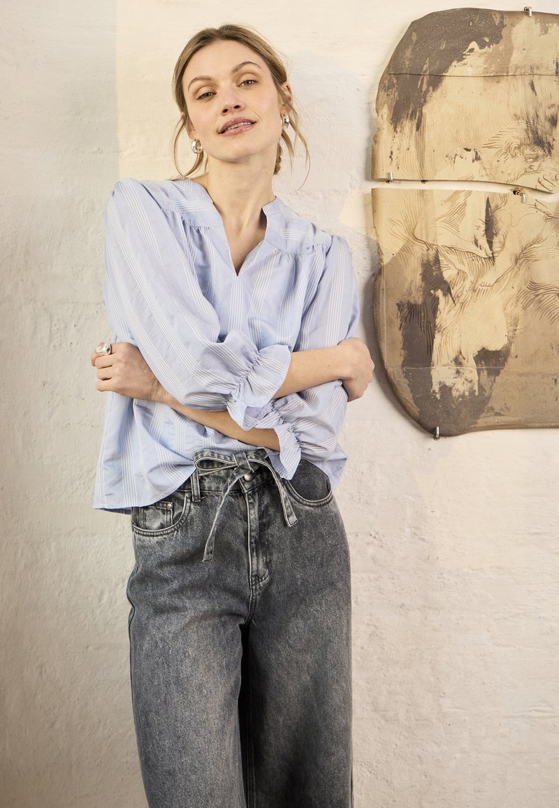 Femme aux cheveux blonds portant un chemisier bleu rayé et un jean gris taille haute, debout les bras croisés près d'un mur texturé et d'une œuvre d'art abstraite monochrome.