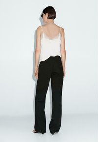 Camisole blanche avec dentelle portée sur un corps avec un pantalon noir à jambes larges. Tissu doux avec une coupe décontractée, vu de dos.