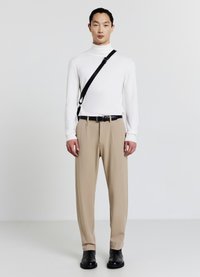 Uomo che indossa un dolcevita bianco, pantaloni beige, cintura nera, borsa a tracolla nera e stivali neri, in piedi contro uno sfondo bianco semplice.