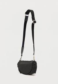 Liten svart skulderveske med justerbar stropp, sølvdetaljer, glidelåslukking øverst og avrundet frontlomme mot en ensfarget lys bakgrunn.