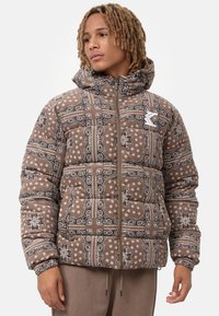 Chaqueta acolchada marrón con un patrón de paisley en blanco y negro, que cuenta con una capucha y un cierre delantero con cremallera. Acentuada con un parche de logo.
