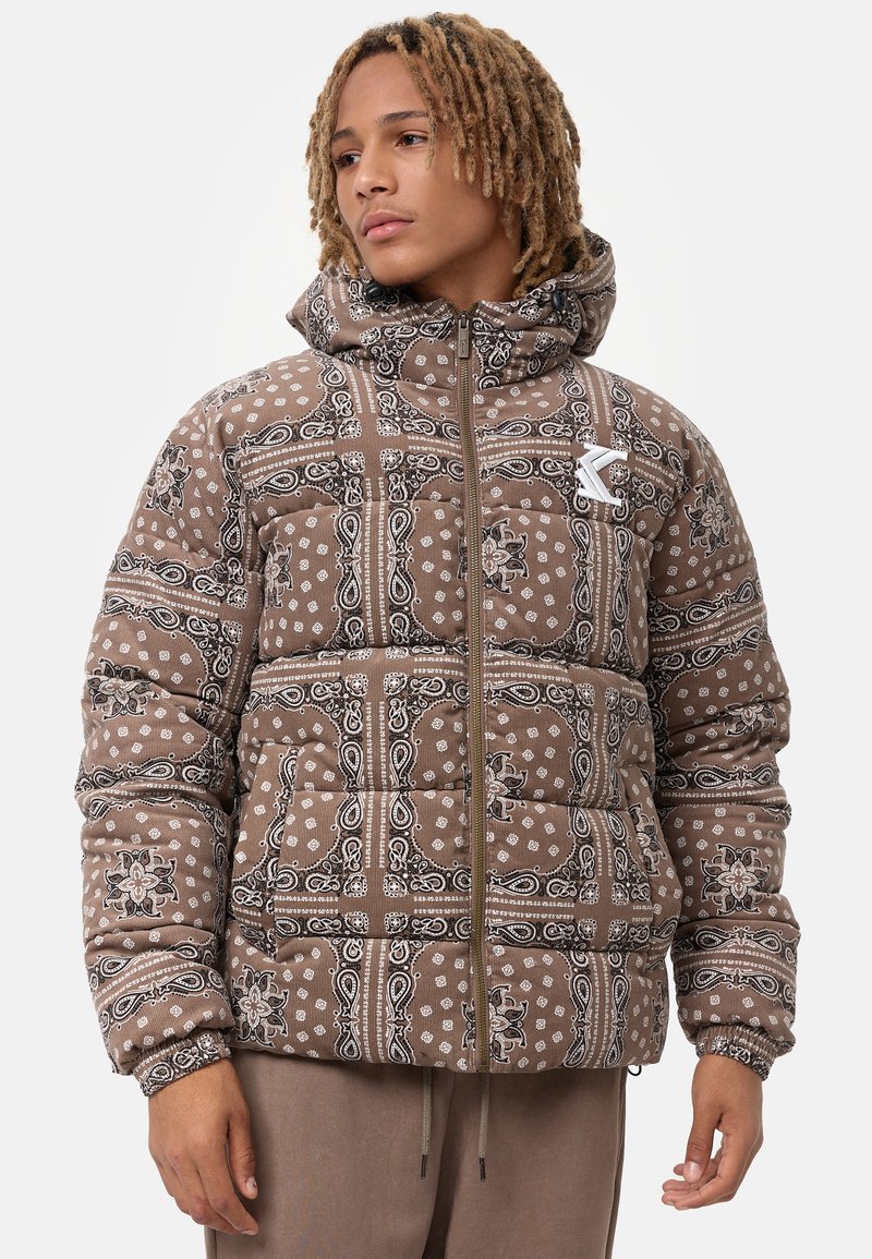 Chaqueta acolchada marrón con un patrón de paisley en blanco y negro, que cuenta con una capucha y un cierre delantero con cremallera. Acentuada con un parche de logo.