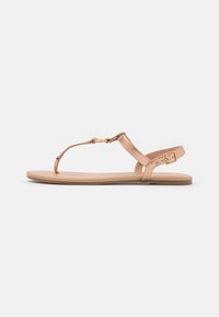 Läskär roséguld sandal med tunna remmar, en guld cirkulär detalj och en justerbar spänne på en beige sula mot en vit bakgrund.
