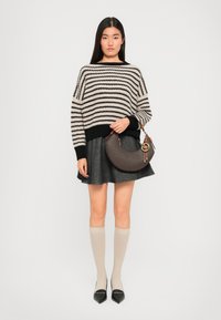 Frau in einem gestreiften schwarzen und beigen Pullover, grauem Rock, beigefarbenen Knietiefen Socken und schwarzen spitzen Schuhen, die eine braune halbmondförmige Handtasche hält.