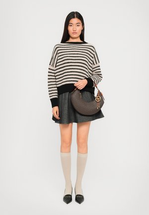 Kvinna iförd en randig svart och beige tröja, grå kjol, beige knähöga strumpor, svarta spetsiga skor och håller en brun halvmåne-handväska.