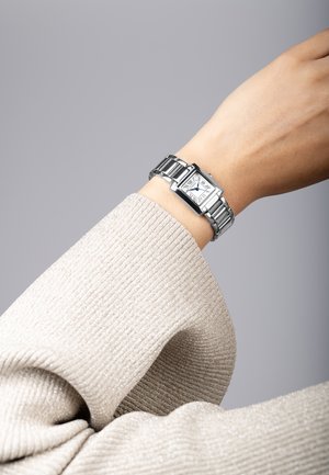 Montre rectangulaire en argent avec un cadran blanc, des aiguilles bleues et un bracelet en métal. Portée sur un poignet portant un pull texturé de couleur claire.