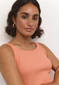 Débardeur côtelé pêche avec un col rond et des bretelles larges. Le mannequin porte des boucles d'oreilles en or et un collier délicat en or.
