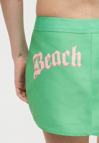 Πράσινη φούστα με ροζ γοτθικό κείμενο "Beach" στην πλάτη, εμφανίζεται στο κάτω μέρος του κορμού και στο χέρι ενός προσώπου με μικρό τατουάζ στον καρπό.