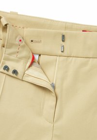 Beżowe spodnie z zapięciem na zamek i guzik, z czerwonymi przeszyciami. Materiał jest gładki, o jednolitej fakturze, bez widocznych wzorów.