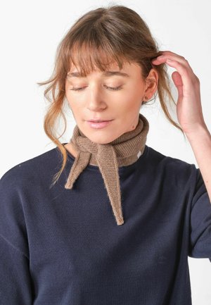 Femeie cu păr castaniu deschis, purtând un pulover bleumarin și un fular bej tricotat legat, mângâindu-și părul cu ochii închiși pe fundal alb.