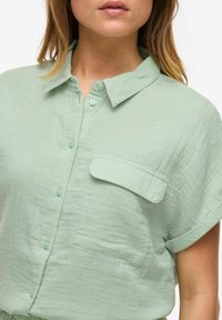 Camicia verde chiaro con bottoni realizzata in tessuto testurizzato, dotata di colletto, maniche corte e taschino sul lato sinistro del petto.