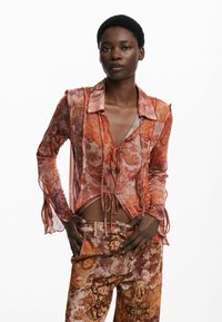 Blusa transparente con estampado floral en cálidos tonos de naranja y rosa, con un lazo en la parte delantera, mangas largas y tela suave y fluida. Combinada con pantalones a juego.