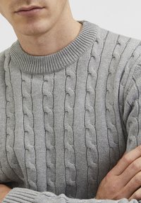 Suéter gris de punto con un patrón de trenzas gruesas, que presenta un cuello redondo acanalado y una tela texturizada. Primer plano del escote y detalles frontales.