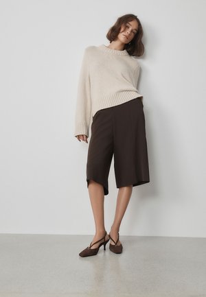 Donna con maglione oversize beige, pantaloncini scuri al ginocchio e scarpe con tacco marroni, in piedi contro un muro bianco con una posa rilassata.