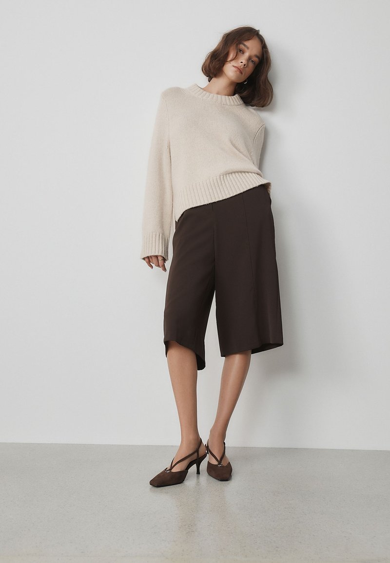 Femme portant un pull oversize beige, un short sombre au niveau des genoux et des chaussures à talons marron, debout contre un mur blanc dans une pose décontractée.