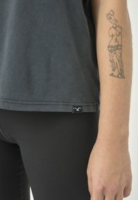 Graues Baumwoll-T-Shirt mit kurzen Ärmeln, seitlichem Etikett und lockerer Passform, kombiniert mit schwarzen Leggings. Sichtbares Tattoo am Unterarm.