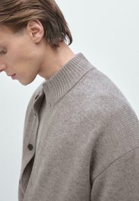 Jasnobrązowy sweter z dzianiny z wysokim prążkowanym kołnierzem i zapięciem na guziki. Gładka faktura i luźny krój. Minimalistyczny design.