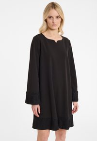 Schwarzes Kleid mit einem dezenten V-Ausschnitt, langen Ärmeln und strukturierten Fransenärmeln, die mit Perlen verziert sind. Glattes Material mit lockerer Passform.