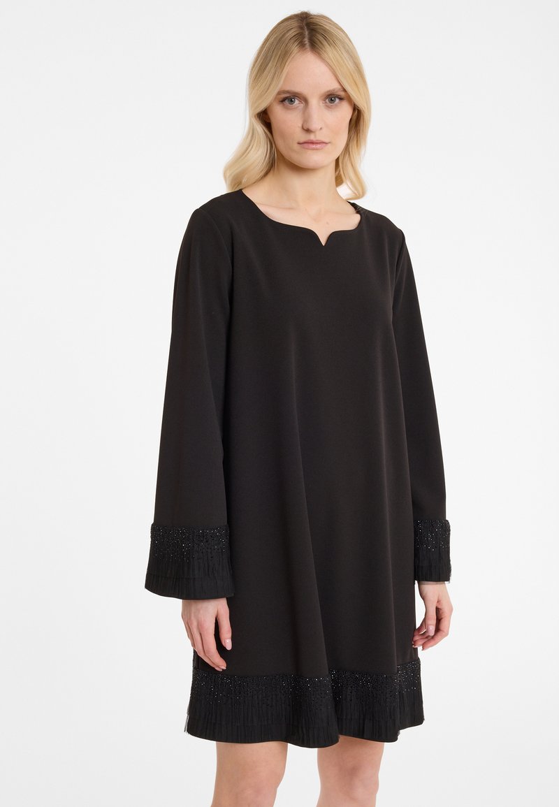 Schwarzes Kleid mit einem dezenten V-Ausschnitt, langen Ärmeln und strukturierten Fransenärmeln, die mit Perlen verziert sind. Glattes Material mit lockerer Passform.