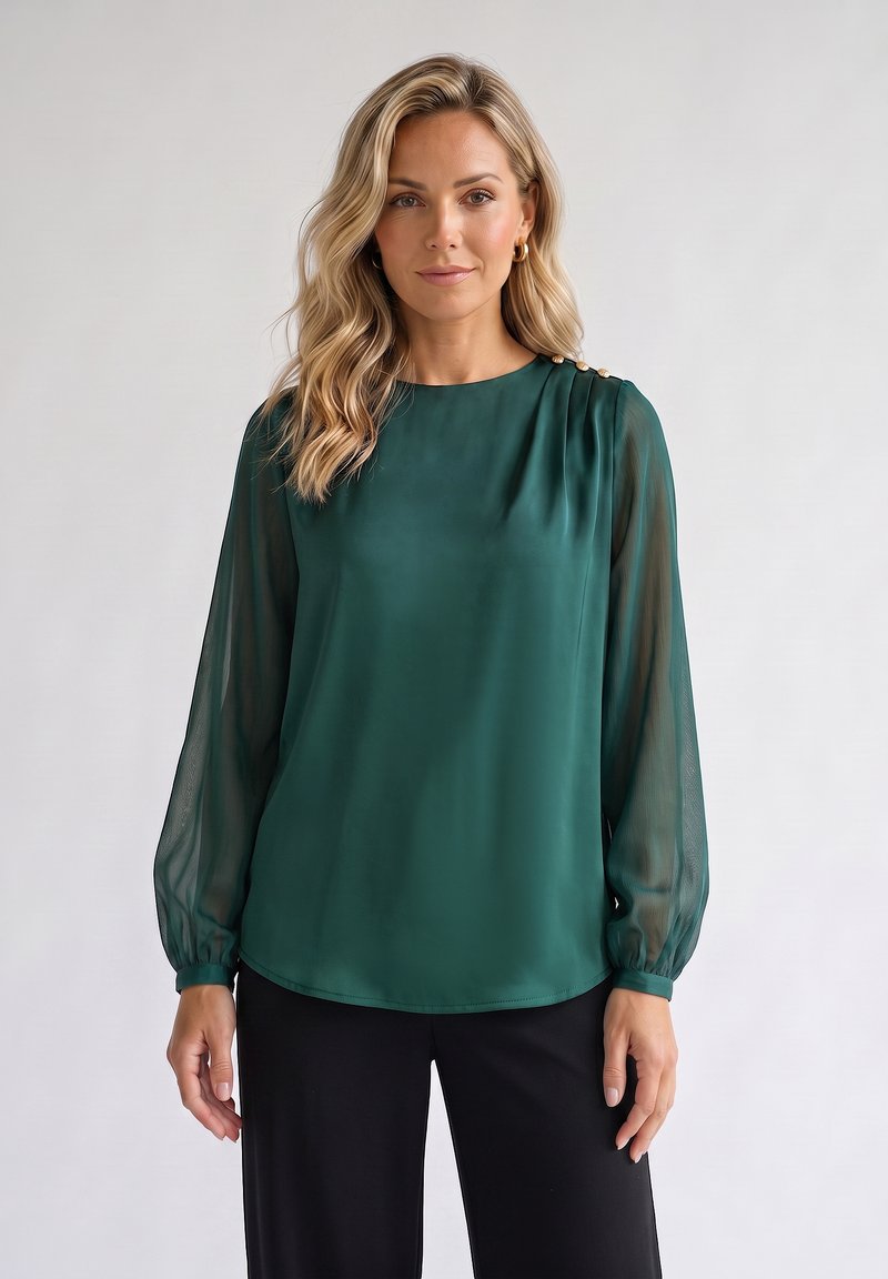 Femme aux cheveux blonds portant un chemisier vert foncé avec des manches longues transparentes et un pantalon noir, debout devant un fond clair uni.