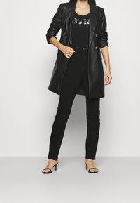 Cappotto doppio petto in ecopelle nera, top aderente nero con dettagli di paillettes, jeans neri slim e sandali neri con tacco.