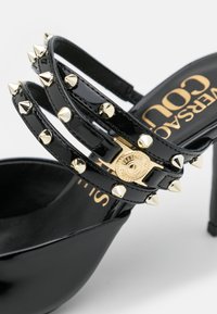 Sandale à talon haut en cuir verni noir avec des clous coniques dorés et une boucle logo Versace dorée sur trois brides.