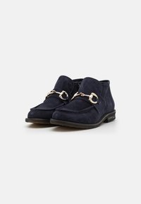 Zapatos de ante azul marino con punta redondeada, correa frontal con herrajes metálicos y tacón bajo. El diseño incluye un borde decorativo cosido.