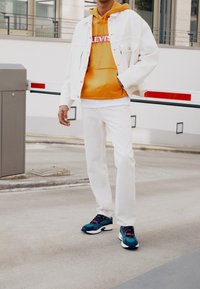 Bílá džínová bunda přes oranžovou mikinu, doplněná bílými džíny a modrými teniskami. Tento outfit obsahuje odvážné barvy a ležérní designové prvky.
