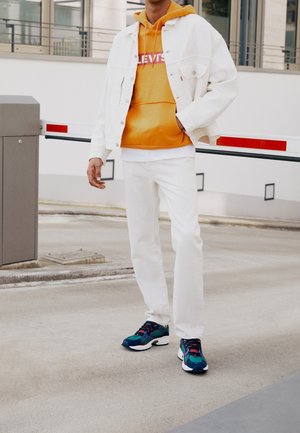 White denim jacket over an orange hoodie, paired with white jeans and blue sneakers. The outfit features bold colors and casual design elements.