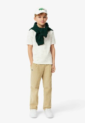 Bambino che indossa una polo bianca a fantasia, un maglione verde annodato attorno al collo, pantaloni di corduroy beige e sneakers bianche, con un cappellino bianco.