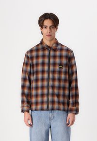 Plaid zip-up jacket in brown, blue, and orange tones. Features a collar, long sleeves, and a logo on the chest. Cotton blend material.