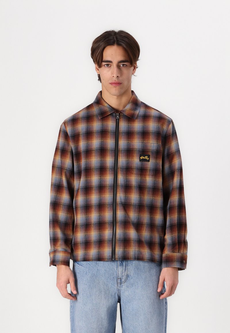 Plaid zip-up jacket in brown, blue, and orange tones. Features a collar, long sleeves, and a logo on the chest. Cotton blend material.