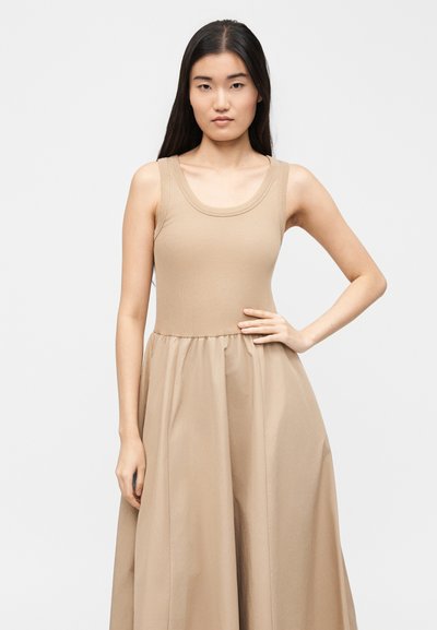 Νεαρή γυναίκα με μακριά σκούρα μαλλιά φοράει αμάνικο μπεζ φόρεμα με εφαρμοστό μπούστο και φουσκωτή φούστα, στέκεται μπροστά σε μονόχρωμο φόντο.