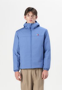 Jachetă puf light blue cu fermoar frontal, glugă ajustabilă, manșete elastice și un mic detaliu cu logo pe piept. Textură de stofă moale.