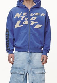 Sudadera azul con cremallera que presenta la inscripción en blanco "NUNCA ES TARDE" en el frente y "PEGADOR" en negro en la manga izquierda. Tiene bolsillos tipo canguro y está hecha de material de algodón.