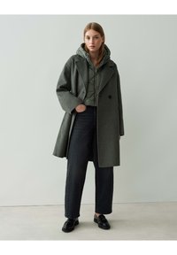 Grau wollener Übermantel über einer grünen gepolsterten Jacke, kombiniert mit dunklen Jeans und schwarzen Schuhen. Der Übermantel hat eine lockere Passform und große Revers.