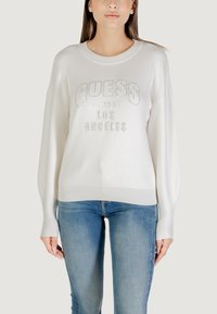 Witte pullover sweatshirt met "GUESS" in zilveren strass-steentjes, ronde hals, verlaagde schouders en geribbelde zoom. G gedragen met blauwe spijkerbroeken.