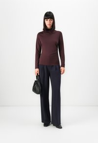 Vrouw met recht zwart haar die een maroon top met lange mouwen, marineblauwe wijde broek, zwarte schoenen draagt en een zwarte handtas vasthoudt.