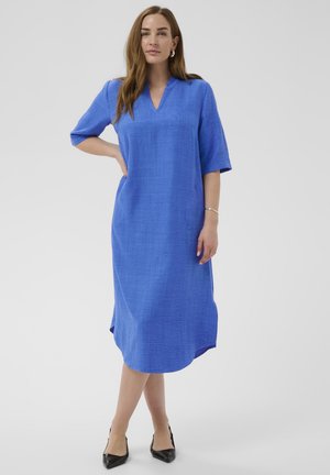 Femme debout, main sur la hanche, portant une robe bleu vif au genou avec des manches mi-longues et des talons pointus noirs sur un fond blanc.