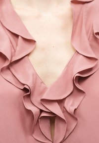 Blouse rose avec des détails à volants superposés le long du décolleté en V profond sur peau claire.
