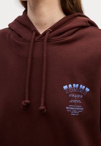 Gros plan d'une personne portant un sweat à capuche marron avec des cordons ajustables et un logo bleu "Tommy Jeans" sur la poitrine.