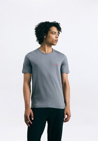 Tricou din bumbac gri, cu decolteu rotund și mâneci scurte, având un mic logo roșu "HUGO" pe partea stângă. Textură uniformă.