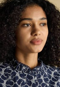 Tumesinine riietus keerukate valgete lillekülvide ja volangkaelusega, mis omab pehmet tekstuuri ja tihedat lõiget.