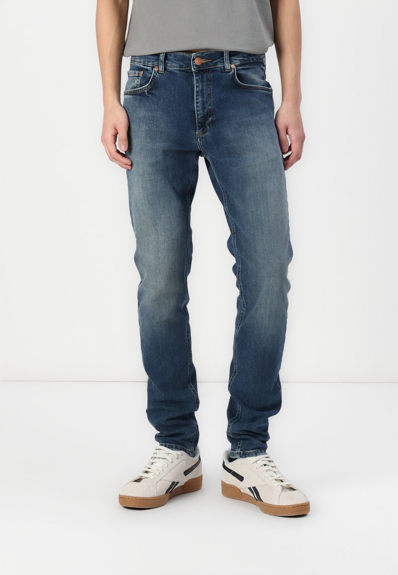 Jeans de denim azul desgastado con un ajuste slim, diseño de cinco bolsillos y herrajes en tono cobre. Combinados con zapatillas de colores claros con acentos negros.