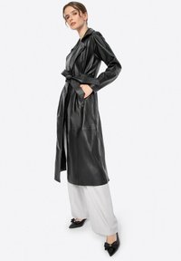 Cappotto trench in pelle nera con vita regolabile, colletto a revers e maniche lunghe, abbinato a pantaloni larghi grigio chiaro e scarpe nere.