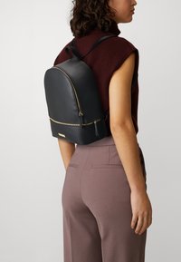 Un sac à dos en cuir noir de forme arrondie, avec une fermeture éclair dorée et des bretelles ajustables, porté sur le dos d'une personne.