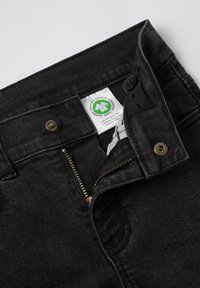 Mustat denim-farkut, joissa on vetoketju ja kultainen vetoketjun osa sekä nappikiinnitys. Sisävyötäröllä näkyvä orgaaninen tekstiililappu.