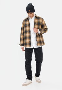 Bruin en zwart geruite flanellen shirt over een witte blouse, zwarte broek en witte sneakers met beige accenten. Zwarte beanie met logo.